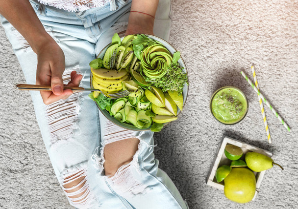 Frau in Jeans mit frischem gesunden grünen Salat mit Avocado, Kiwi, Apfel, Gurke, Birne, Gemüse und Sesam auf hellem Hintergrund. Gesundes Essen, sauberes Essen, Buddha-Salat, Draufsicht, Entgiftung - Foto, Bild