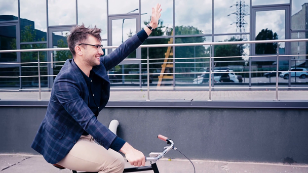 hombre feliz en gafas saludando la mano mientras monta en bicicleta cerca del edificio
  - Imágenes, Vídeo