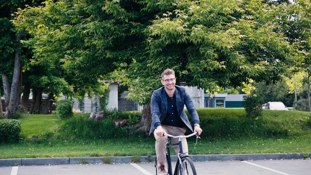 happy man in glasses riding bicycle on asphalt near green trees - Felvétel, videó