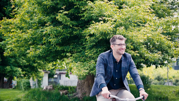 homme heureux dans des lunettes agitant la main tout en faisant du vélo près des arbres - Séquence, vidéo