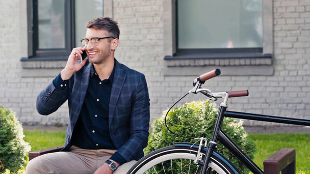 felice uomo in bicchieri agitando la mano e parlando su smartphone vicino alla bici  - Filmati, video