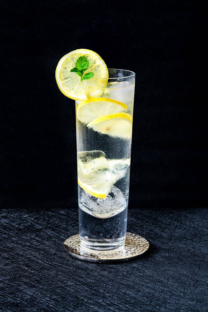 Water with lemon and ice cubes garnished with a slice of lemon and mint on dark background - refreshing cold non alcoholic drink - 写真・画像