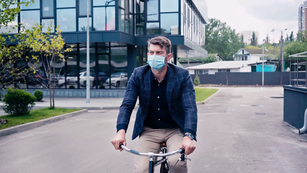 uomo in maschera medica in sella alla bicicletta su asfalto vicino edificio  - Filmati, video