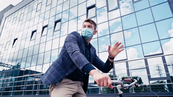 homme d'affaires en masque médical équitation vélo et agitant la main près du bâtiment  - Séquence, vidéo