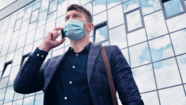 vista basso angolo di uomo d'affari in maschera medica parlando su smartphone vicino edificio - Filmati, video