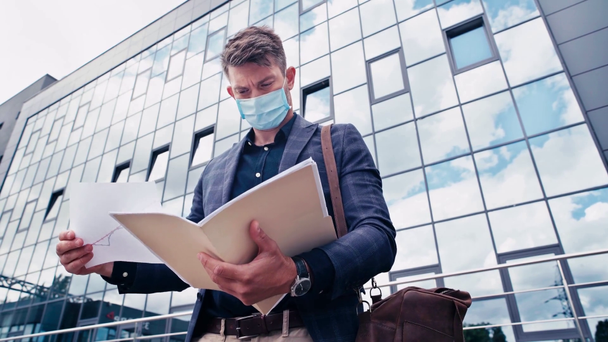 χαμηλή γωνία άποψη του επιχειρηματία σε ιατρική μάσκα κρατώντας φάκελο κοντά στο κτίριο - Πλάνα, βίντεο