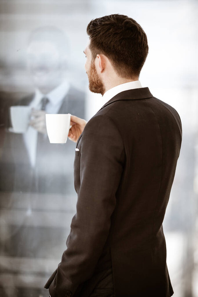 Geschäftskonzept. Erfolgreicher Jungunternehmer zurück am Arbeitsplatz. Manager steht im Büro und trinkt glücklich Kaffee aus Tasse. Mann im Anzug drinnen auf Glasfensterhintergrund. - Foto, Bild