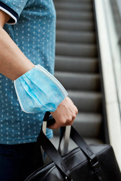 closeup of a young man on a scalator, in casual wear, carrying a travel bag in one hand and a suitcase in the other hand, wearing a surgical mask in his arm while is not wearing it in his face - Foto, Bild