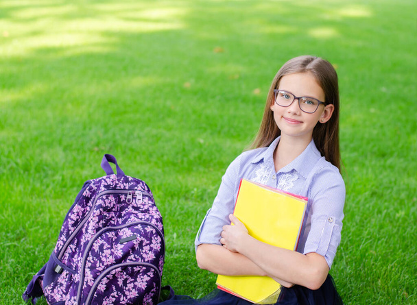 Back to school. Education concept. Cute smiling schoolgirl in glasses sitting on grass. Happy little girl child holding the books - Foto, Imagem