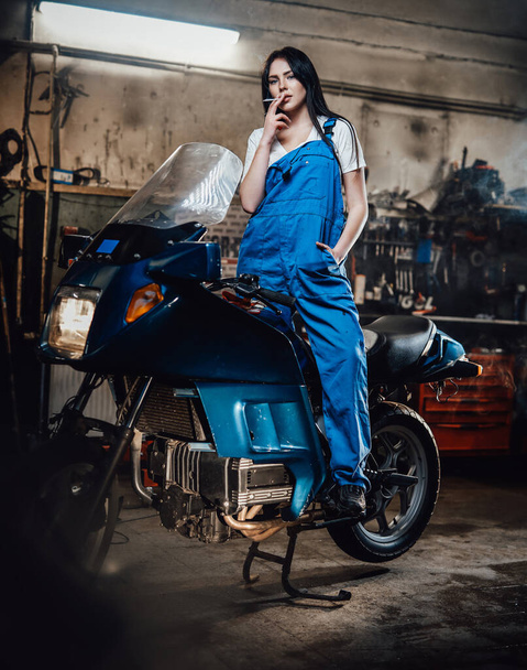 Female mechanic relaxing smoking a cigarette while standing on sportbike in garage or workshop - 写真・画像