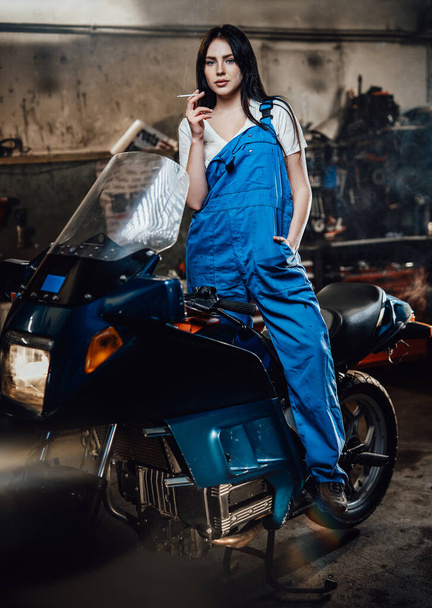 Samice mechanik relaxační kouření cigarety, zatímco stojí na sportovním kole v garáži nebo dílně - Fotografie, Obrázek