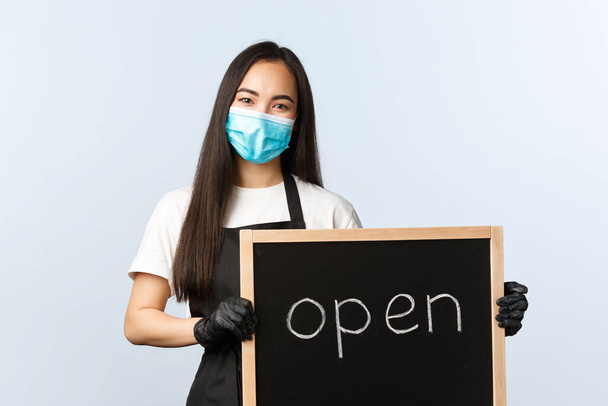 Small business, covid-19 pandemic, preventing virus and employees concept. Smiling cute asian female employee, cafe or store worker holding open sign, invite visitors - Фото, изображение
