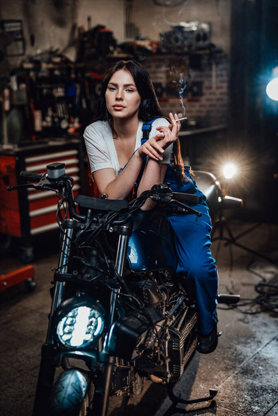 Hermosa morena motorista o mecánico relajante fumar un cigarrillo en el garaje o taller
 - Foto, Imagen