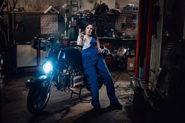 Female mechanic relaxing smoking a cigarette while leaning on custom bobber in garage or workshop - Foto, Bild