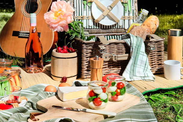 Vintage picnic basket, hamper with baguette and lemonade outdoors on a grass with cheese, mozzarella, tomatoes, cherries, vine. Guitar on straw blanket and stripy towels. Zero waste picnic al fresco. - Fotoğraf, Görsel