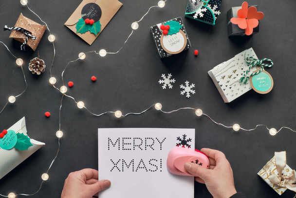 Text Merry Christmas on perforated paper. Presents wrapped in gift boxes with tags. Hands holding greeting text. Flat lay, top view with light garland, green alarm, snowflakes on black paper. - Photo, Image