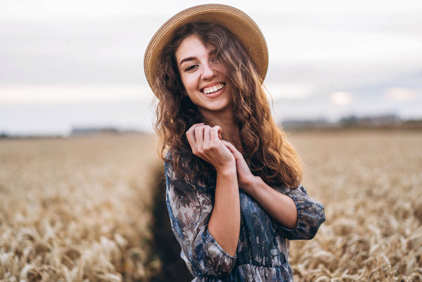 Closeup portrait of a beautiful young woman with curly hair. Woman in dress and hat standing in wheat field. - Valokuva, kuva