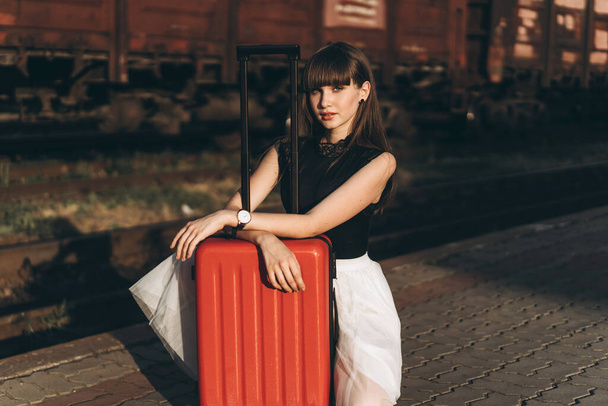 Female brunette traveler with red suitcase in white skirt waiting for a train on raiway station - Fotoğraf, Görsel