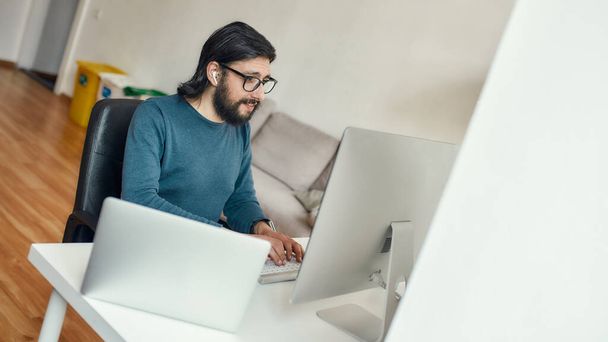 Young caucasian concentrated bearded man sitting at his workplace and working with computer remotely, working from home. Freelance, home office - Foto, immagini