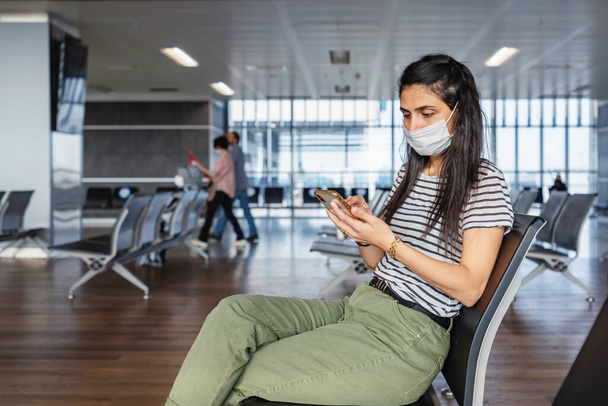 Young woman protection mask is sitting in a waiting hall and looking to her mobile phone at the airport. Corona virus outbreaking. - Фото, изображение