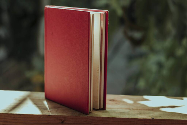 A closeup of a book with a red cover on a wooden fence in a garden under the sunlight - Фото, зображення
