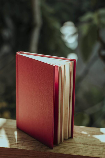 A vertical shot of a book with a red cover on a wooden fence in a garden under the sunlight - Fotó, kép