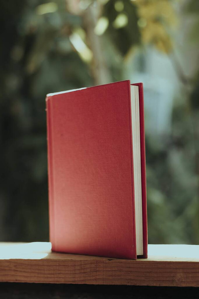 A vertical shot of a book with a red cover on a wooden fence in a garden under the sunlight - 写真・画像