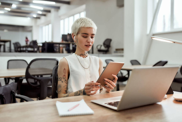 Talking online. Young blonde tattooed business woman in wireless earphones using her touchpad, having skype call with client while sitting at her working place in the office - Foto, Imagen
