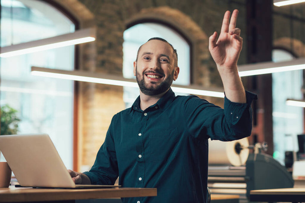 Positive young employee sitting at the table with a laptop and smiling while putting hand up and waving to a colleague - Foto, imagen