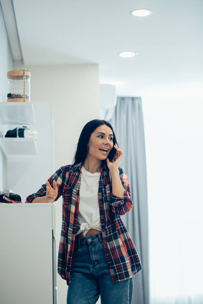 Attractive young woman standing in the room near the chest of drawers and smiling while having a phone talk - Foto, Bild
