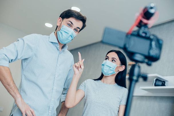 Pohledný muž hledící do kamery, zatímco mladá dáma ukazuje na lékařskou masku na tváři. Koncept koronaviru - Fotografie, Obrázek