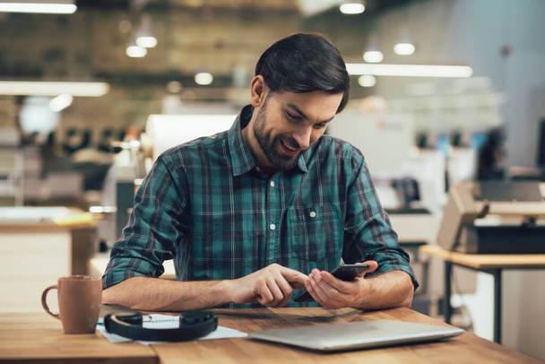 Positive young man in checkered shirt sitting at the office table with coffee and headphones and smiling while looking at the screen of a gadget - 写真・画像