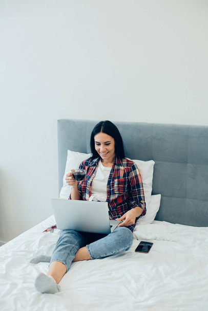 Cheerful lady in casual clothes sitting on the bed with a cup of coffee and smiling while using a modern laptop - Foto, Imagem