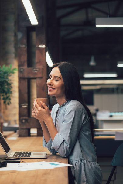 Pleased pretty lady holding a mug and smiling with closed eyes while sitting at the office table - Photo, image