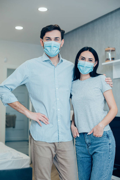 Wearing medical masks at home. Attractive young man hugging his wife by the shoulder - Foto, Imagem