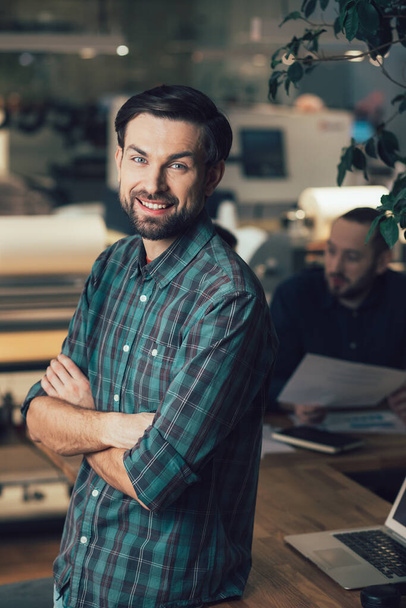 Positive attractive employee standing with his arms crossed next to the table and smiling - Foto, Bild
