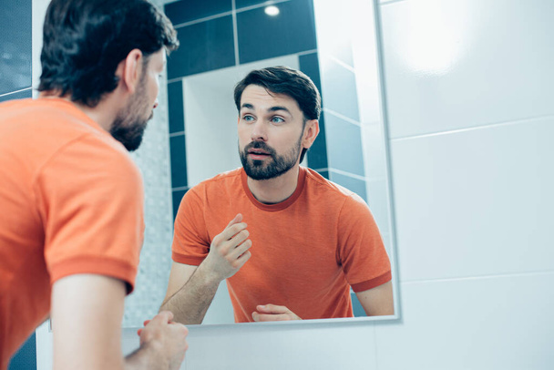 Calma joven atractivo en naranja camiseta de pie en el baño delante de un espejo y mirando a su reflejo
 - Foto, imagen