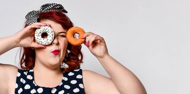 Portrait of beautiful cheerful fat plus size woman pin-up wearing a polka-dot dress isolated over light background, eating a donut - Photo, image
