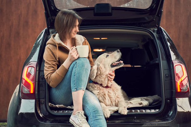 Happy girl with mug in hand with dog sitting in open trunk of black car. - Foto, afbeelding