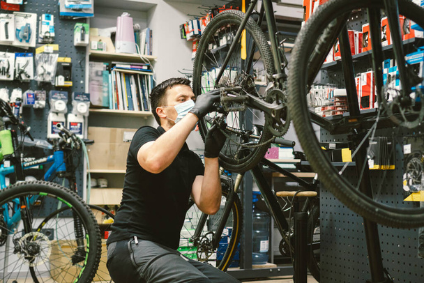 Young man working in a biking repair shop under quarantine and pandemic coronavirus in a protective mask on the face. Bicycle Mechanic Repair Bike, Cycle Workshop in medical mask covid 19 - Foto, Imagen