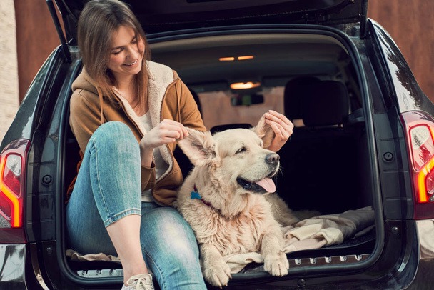 Smiling woman with dog sitting in open trunk of black car - Fotó, kép