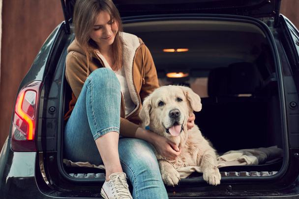 Smiling woman with golden retriever sit in open trunk of black car - Valokuva, kuva