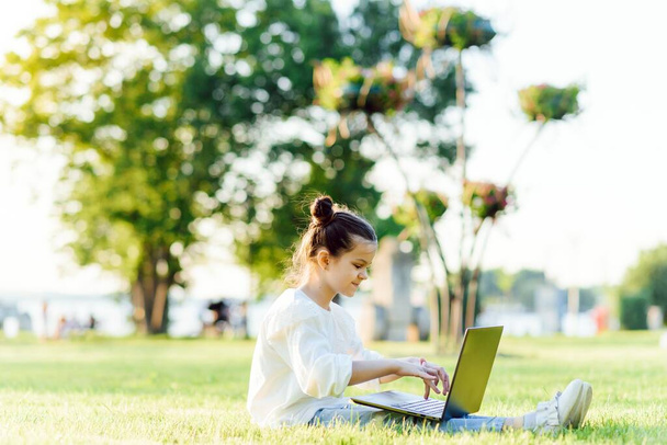 Little girl sitting in the park and working with laptop. Education, lifestyle, technology concept - Foto, afbeelding