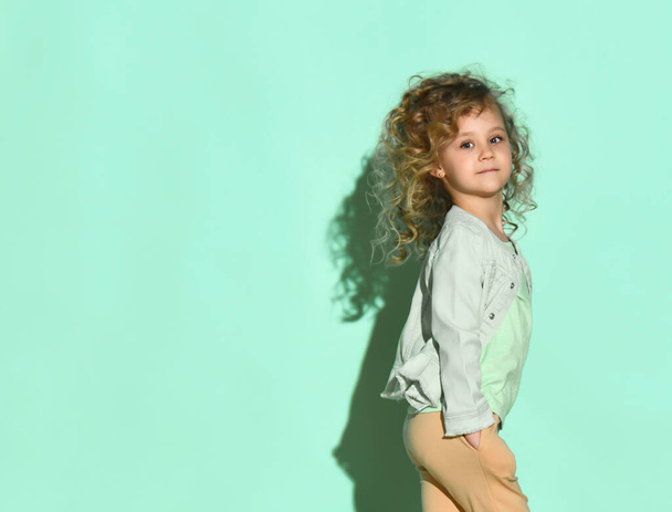 Curly baby in a T-shirt, denim jacket, beige pants. She is smiling standing sideways on a turquoise background. - Photo, Image