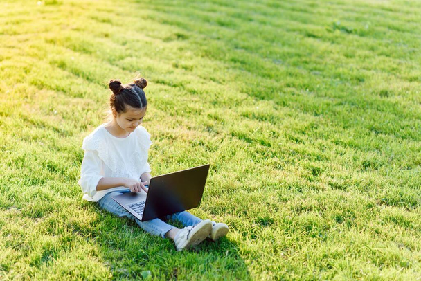 Little girl sitting in the park and working with laptop. Education, lifestyle, technology concept - Photo, image