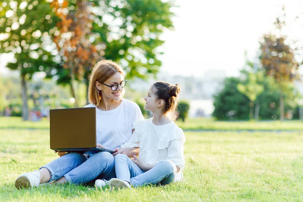 mom and daughter playing in the laptop outdoors, laughing and enjoying the summer sun on the green grass in the park, family outdoors in the Park - Фото, зображення