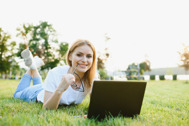 Beautiful and happy young woman lying on the grass surrounded by technology devices - Fotó, kép