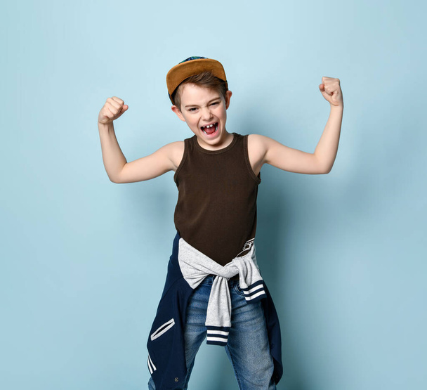 Teenage male in cap and casual stylish clothes. He clenched his fists showing muscles and screaming, posing on blue background - Фото, зображення