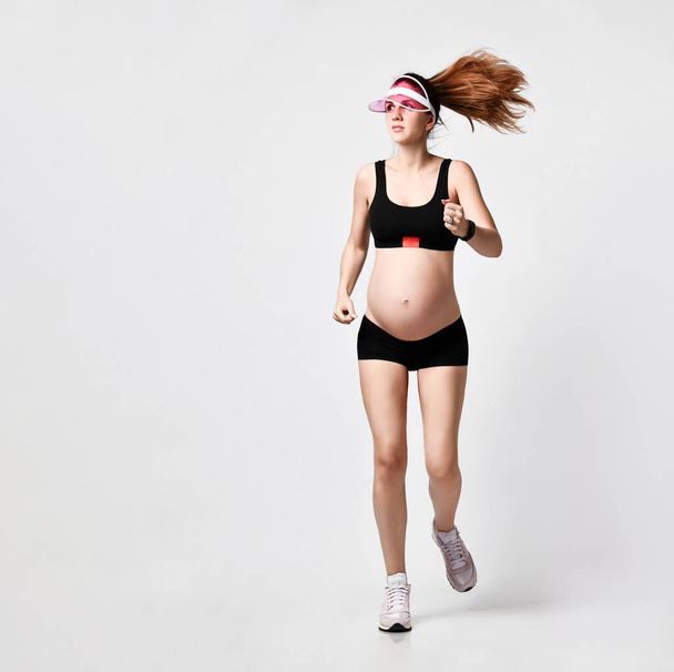 Pregnant lady in sun visor, black sport shorts and top, pink sneakers. She is smiling while jogging isolated on white. Full-length - Photo, image