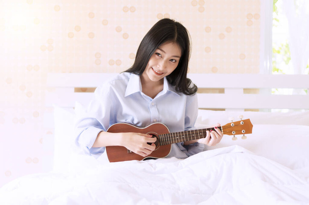 Cheerful smiling young woman playing Ukulele relax on the white bed in the morning. Joyful girl play music with ukulele in bedroom looking at something. - Photo, Image